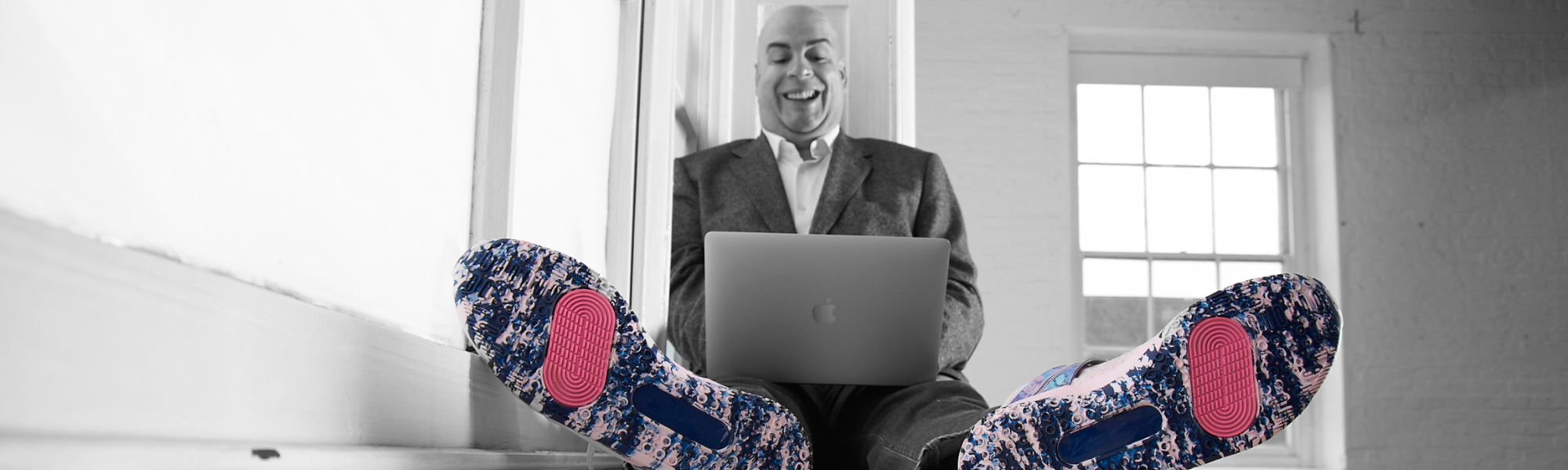 Man sits with his legs stretched out and a laptop computer balanced on them. The soles of his shoes are facing the camera and they have a pattern on them which is pink in places. 