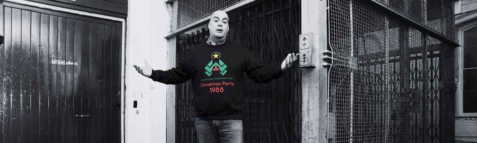 A man stands by a cage elevator wearing sweatshirt with the wording 'Nakatomi Corporation Christmas Party 1988'.