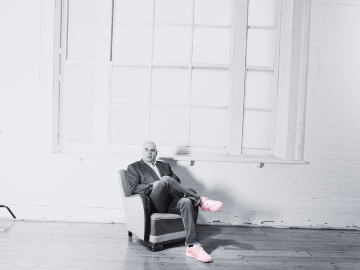 Richard Liverman is wearing jeans, a suit jacket and pink trainers. He sits in front of a large window.